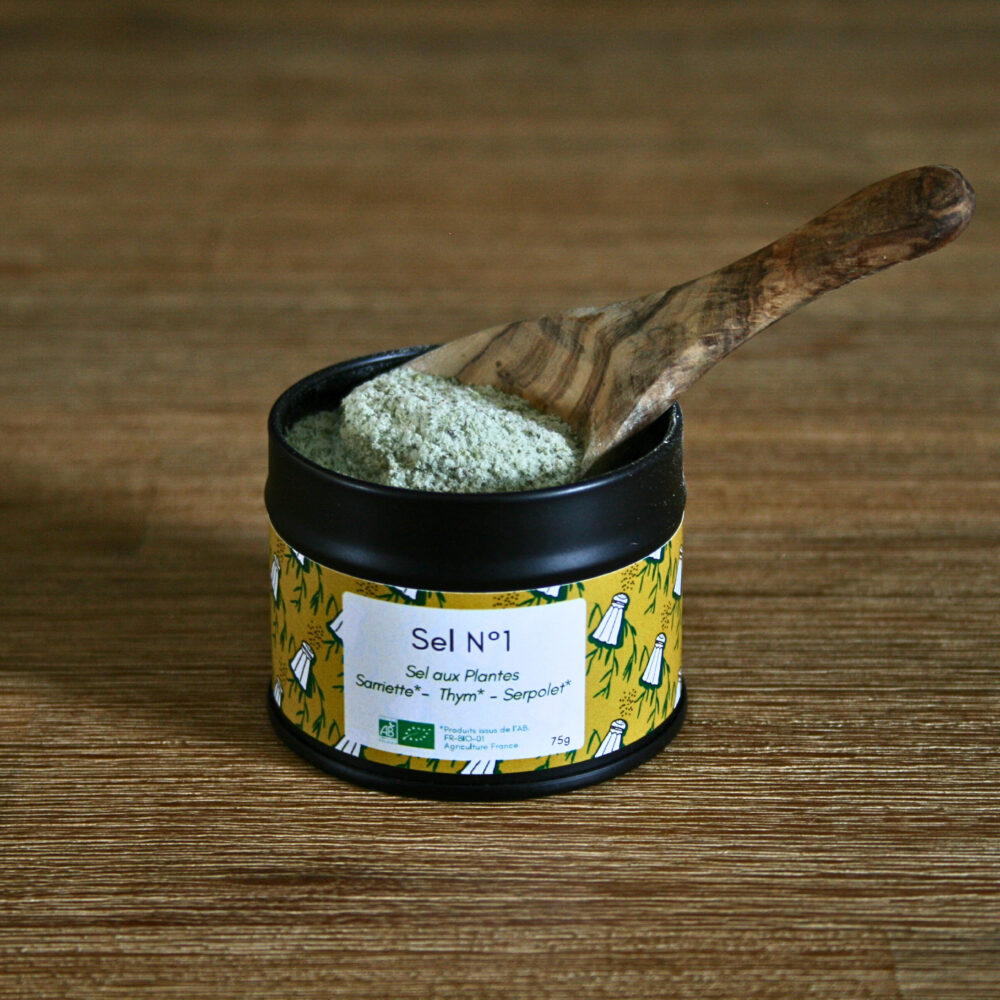 Pot en métal noir ouvert contenant du sel aromatique au thym, sarriette et serpolet, avec une cuillère en bois remplie de sel, posé sur une table en bois rustique.