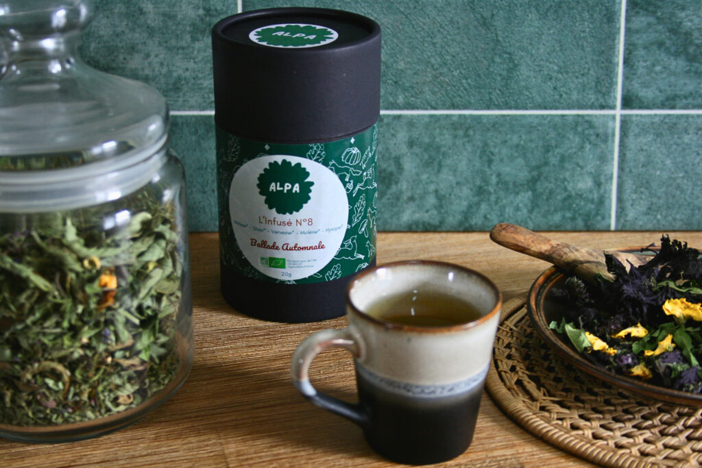 Infusé N°8 – Ballade Automnale dans un pot en kraft noir, posé sur le plan de travail d’une cuisine, accompagné de ses ingrédients dans un bocal en verre et d’une petite tasse de tisane.