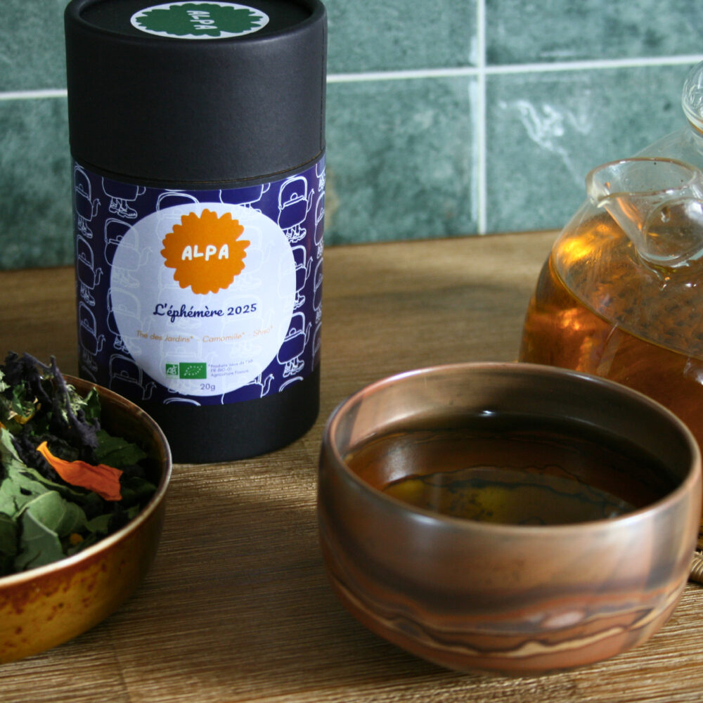 Photo d’un pot en kraft noir contenant l’infusé L’éphémère 2025, à côté un petit contenant avec la composition de l’infusion, une tasse de tisane et une théière sur une table en bois.