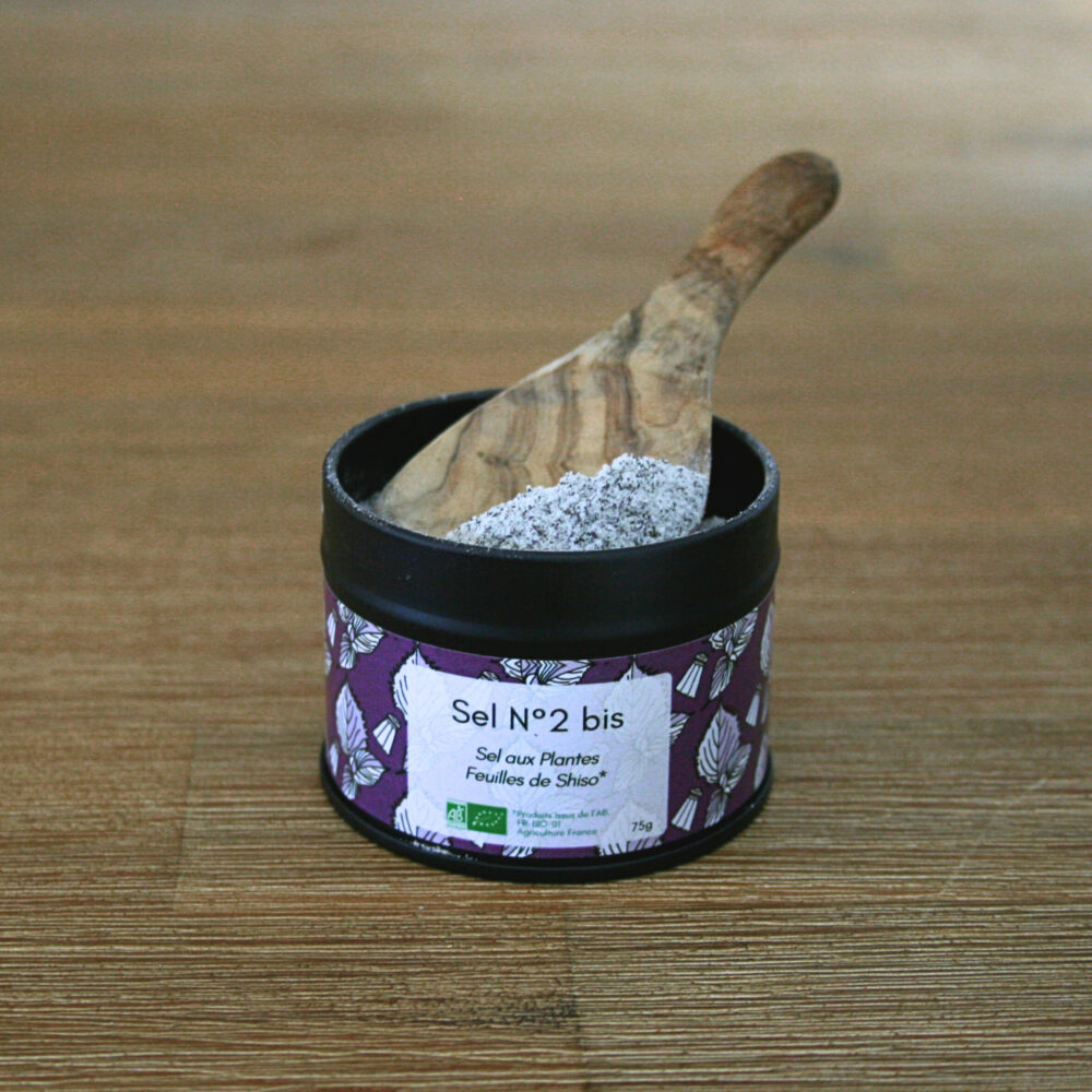 Pot en métal noir ouvert, avec une cuillère en bois remplie de sel moulu et de feuilles de shiso, posé sur une table en bois rustique.