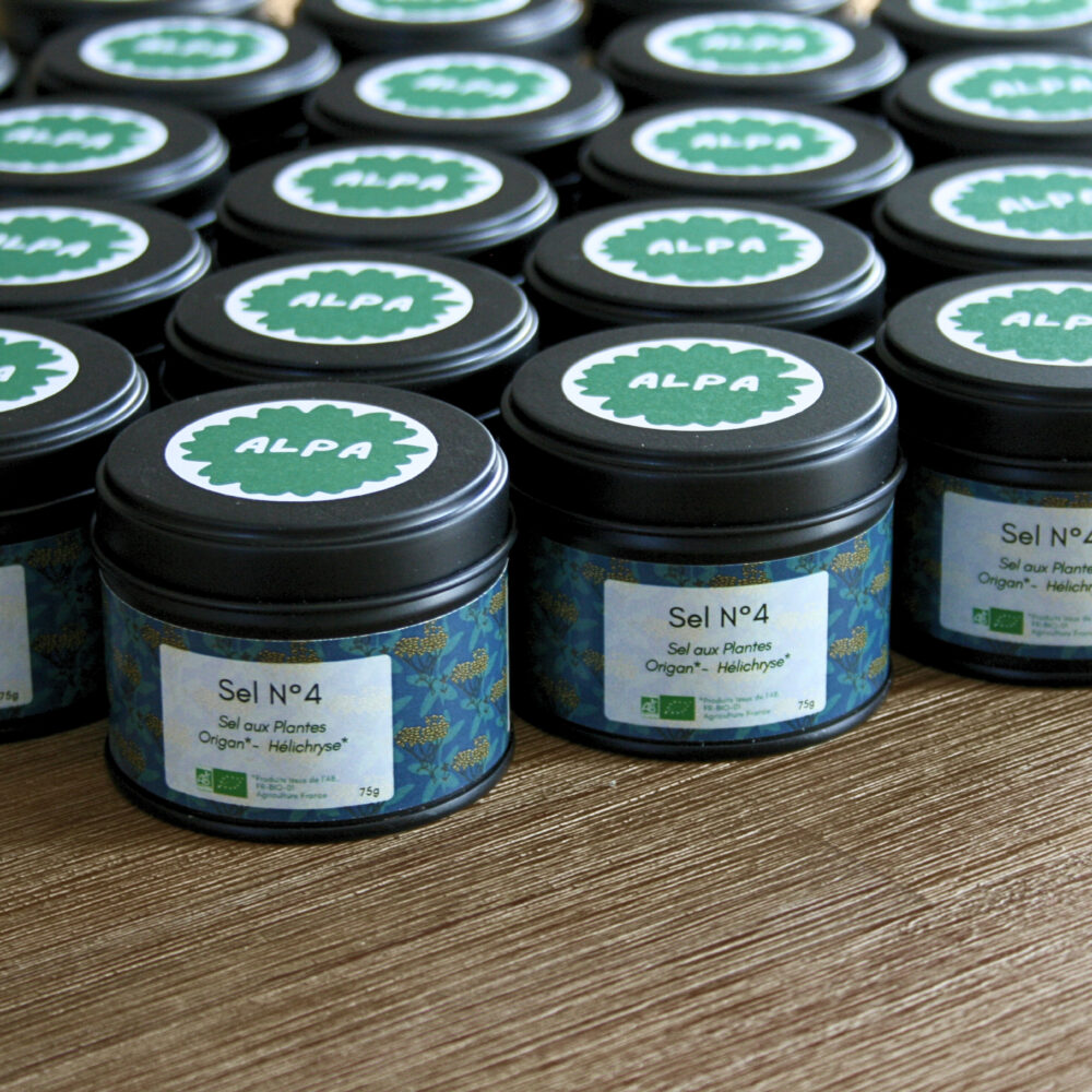 Plusieurs pots en métal noir du Sel N°4 à l’origan et à l’hélichryse, alignés côte à côte sur un plan de travail en bois, évoquant une ambiance artisanale.